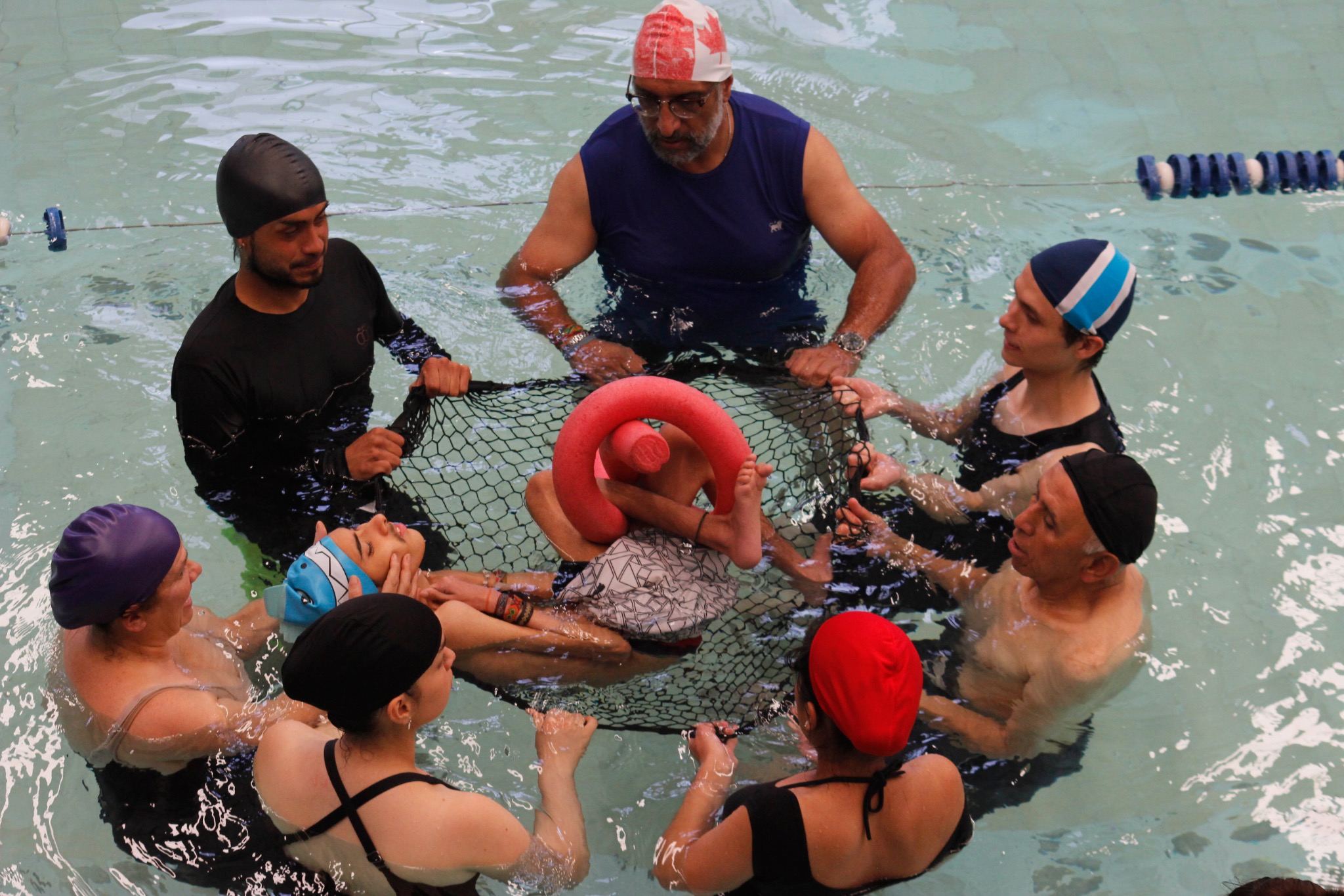 Equipo humano en una piscina sostiene una red alrededor de un participante durante una actividad de acompañamiento colectivo.